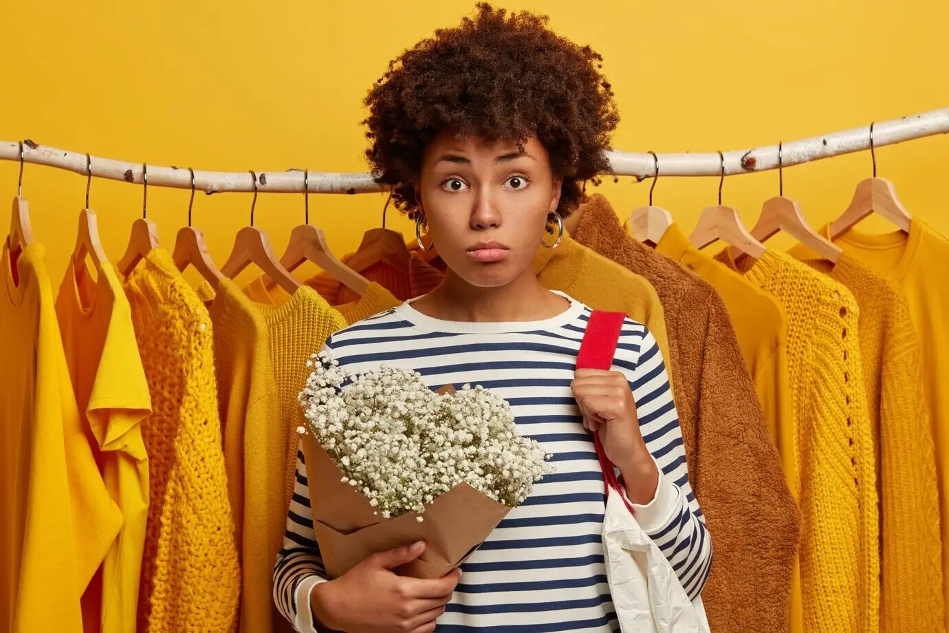 Innenaufnahme einer traurigen, lockigen Frau, die einen gestreiften Matrosenpullover trägt, einen Blumenstrauß hält und eine Tasche auf der Schulter trägt.