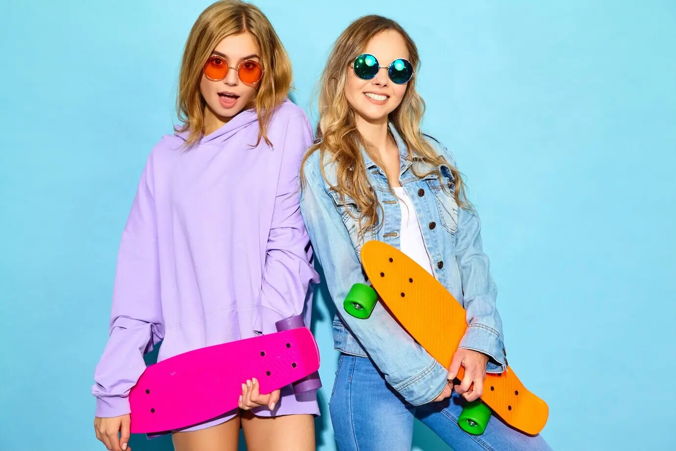 Zwei junge, stylische, lächelnde, blonde Frauen mit Penny-Skateboards. Models in sommerlicher Hipster-Sportkleidung posieren in der Nähe einer blauen Wand. Gut gelaunte Frauen drehen durch.